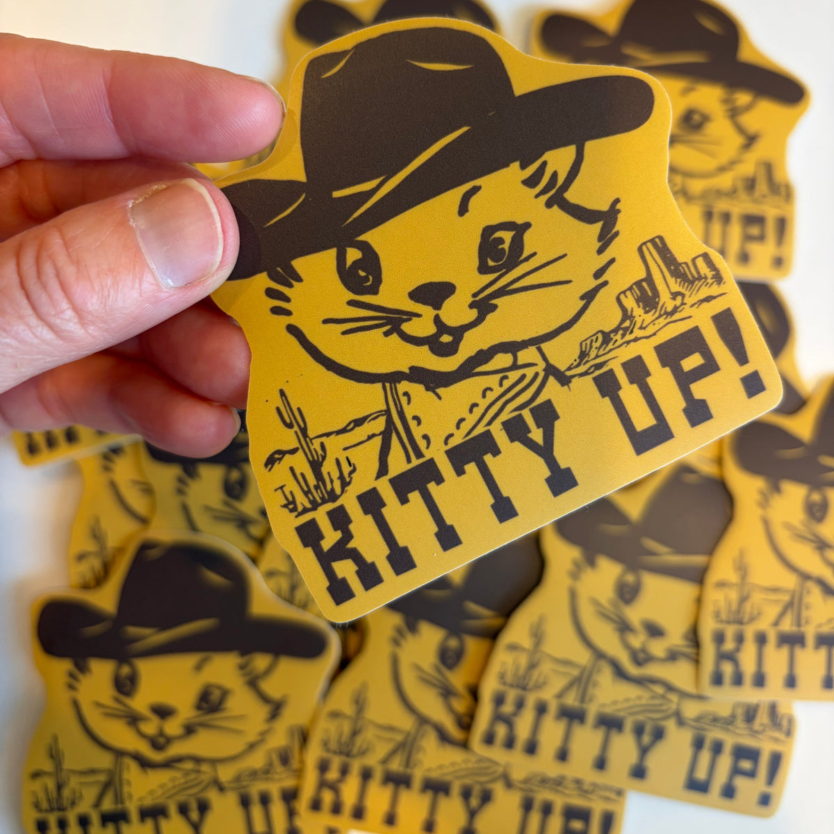 A hand holds a yellow die-cut sticker featuring a cartoon cat in a black cowboy hat with the text 'KITTY UP!' on a white background, with more matching stickers scattered behind it.