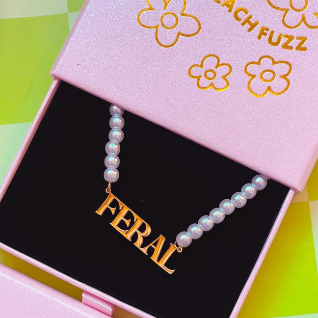 A necklace with the word 'FERAL' written in gold letters, displayed in a pink box with a black interior and flower prints on the lid.