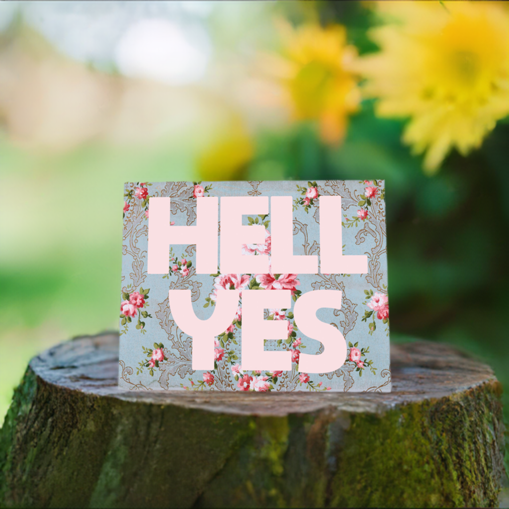 Decorative sign with 'HELL YES' on a floral pattern, placed on a tree stump with flowers in the background.