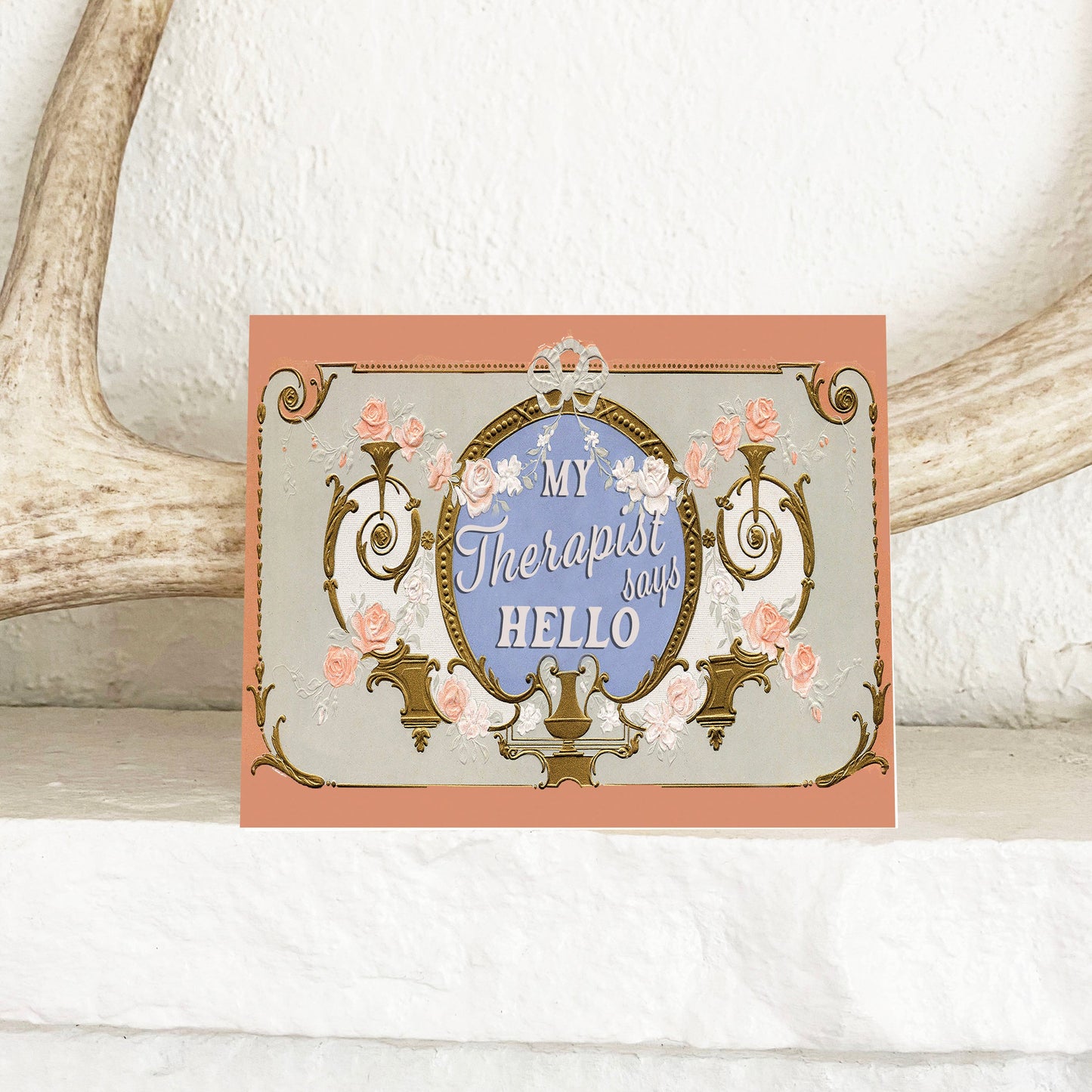 A greeting card with a vintage floral motif and the text 'My Therapist Says Hello' displayed on a white surface, with antique decor in the background.