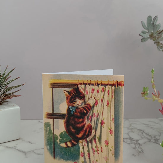 Greeting card with a vintage image of a brown and orange stripped kitty that has climbed up floral drapes covering a window. Greeting says, "Oh fuck." Color pallet is vintage yellows, cream, blues, and pinks. Blank inside. 