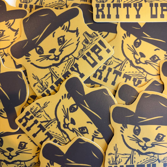 A pile of yellow and black die-cut stickers featuring a cartoon cat wearing a cowboy hat with the text 'KITTY UP!'.