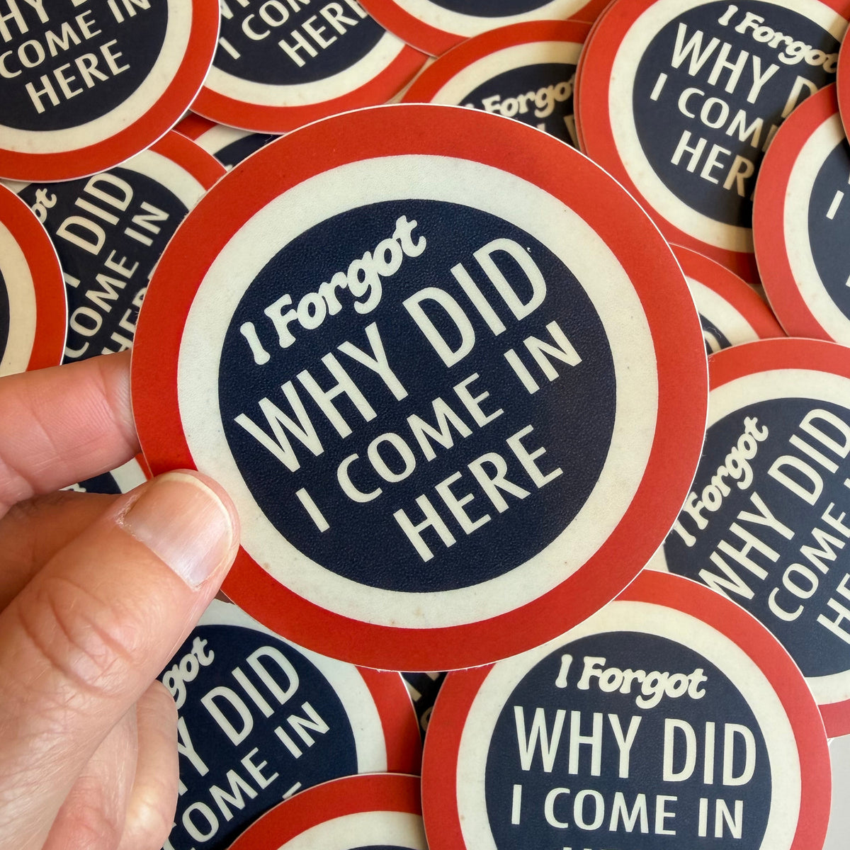 A hand holds a round sticker with the text 'I Forgot WHY DID I COME IN HERE' in white on a navy blue background, surrounded by a red ring and a white inner ring. Multiple identical stickers are scattered in the background.