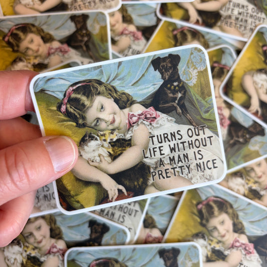 A hand holds a rectangular printed card featuring a vintage-style illustration of a young girl with a cat and a dog, with the text 'TURNS OUT LIFE WITHOUT A MAN IS PRETTY NICE.' Multiple similar cards are scattered in the background.