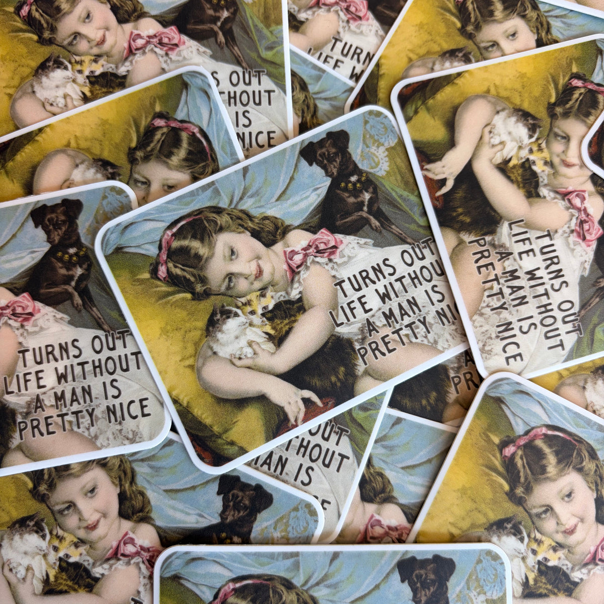 A pile of vintage-style illustrated cards or prints featuring a young girl with a dog, overlaid with the phrase 'TURNS OUT LIFE WITHOUT A MAN IS PRETTY NICE.' The product appears to be the printed cards themselves rather than a lifestyle scene, so this fits Product/Packaging.