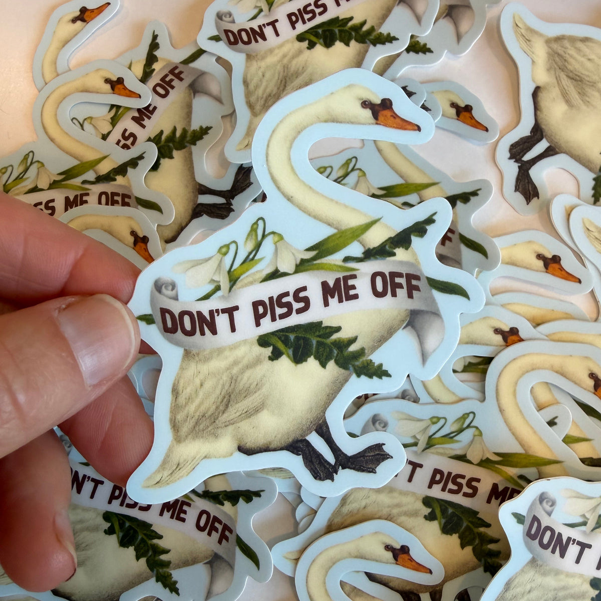 A hand holds a die-cut sticker featuring a white swan with green foliage and the text 'DON'T PISS ME OFF' in brown, surrounded by a pile of identical stickers.