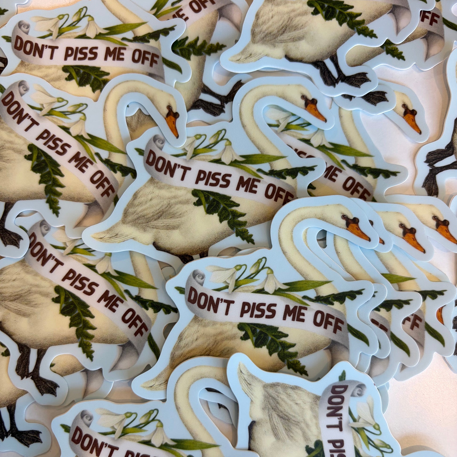 A pile of die-cut stickers featuring a white swan holding a banner that reads 'DON'T PISS ME OFF' in brown text, with green foliage details and a light blue outline.