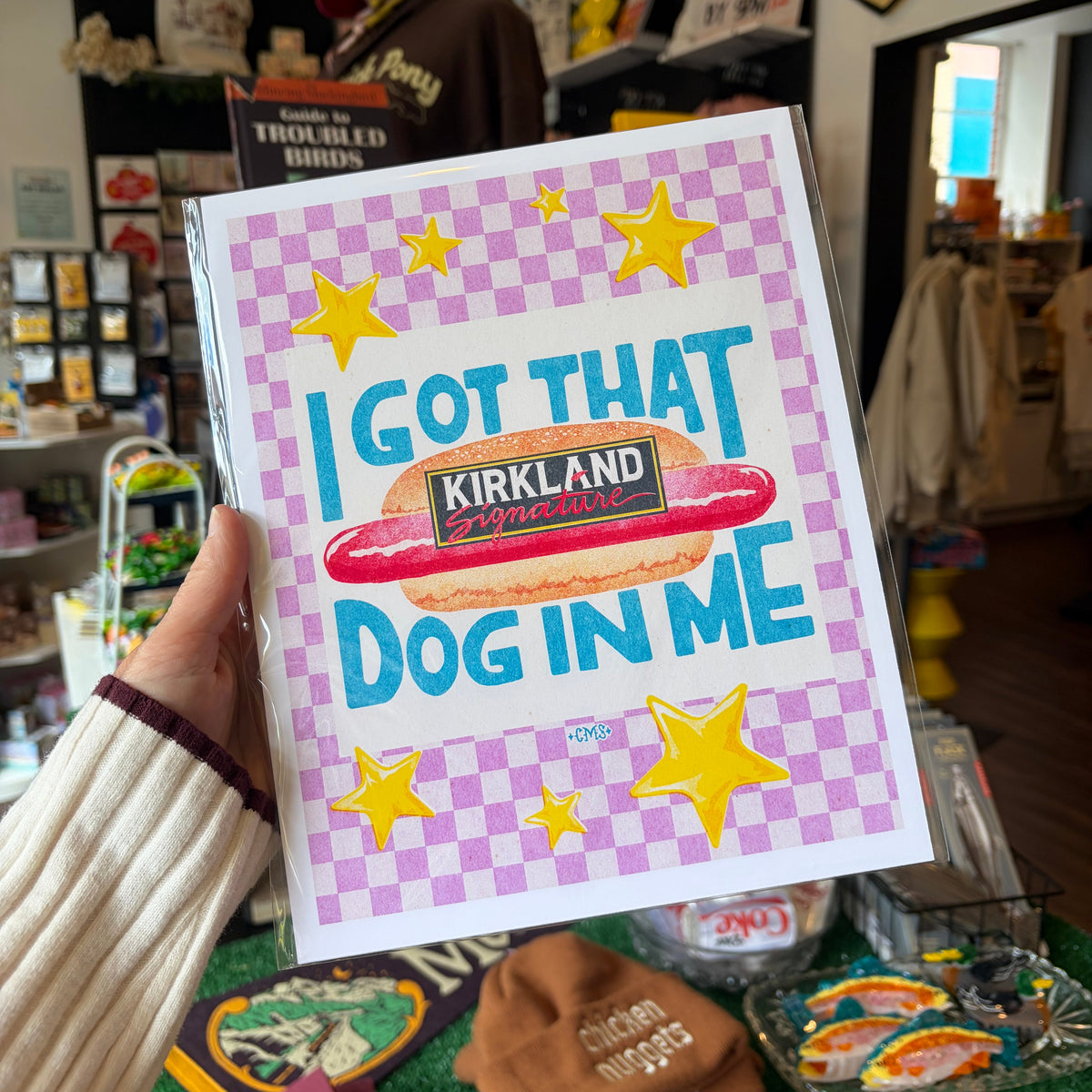 A hand holds a square art print featuring a pink and lavender checkerboard background, yellow stars, and a hot dog with the text 'I GOT THAT KIRKLAND Brand Dog IN ME'. The background shows a busy retail store interior with shelves, merchandise, and a counter.