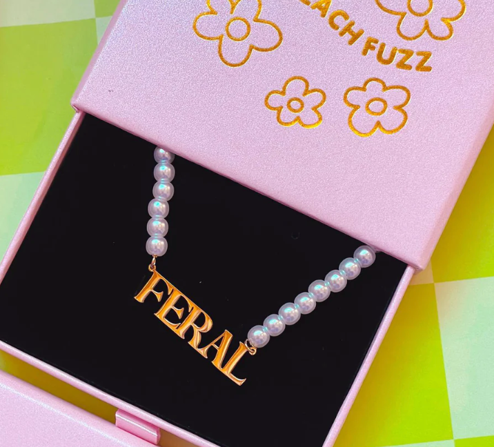 A necklace with the word 'FERAL' written in gold letters, displayed in a pink box with a black interior and flower prints on the lid.
