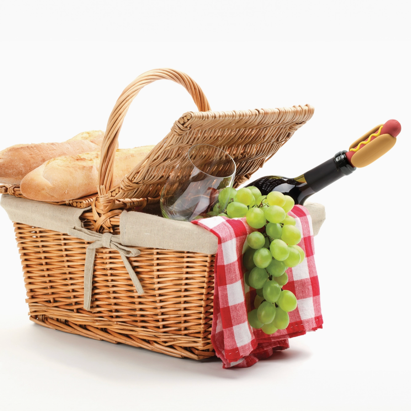 A wicker picnic basket with a fabric liner and ribbon, filled with bread, green grapes, a wine bottle, and a hot dog, photographed on a clean white background.