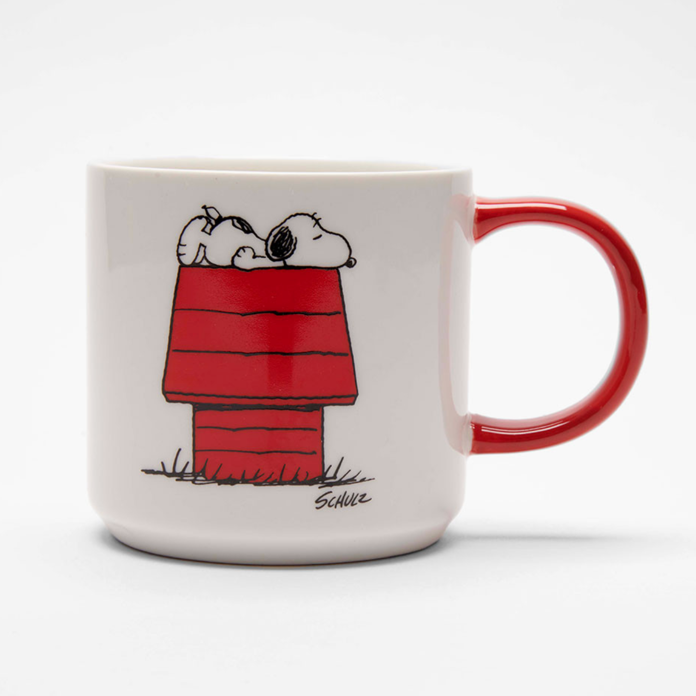 A white ceramic mug featuring the classic Peanuts Snoopy in his red doghouse illustration, with a glossy red handle, photographed on a clean white background.