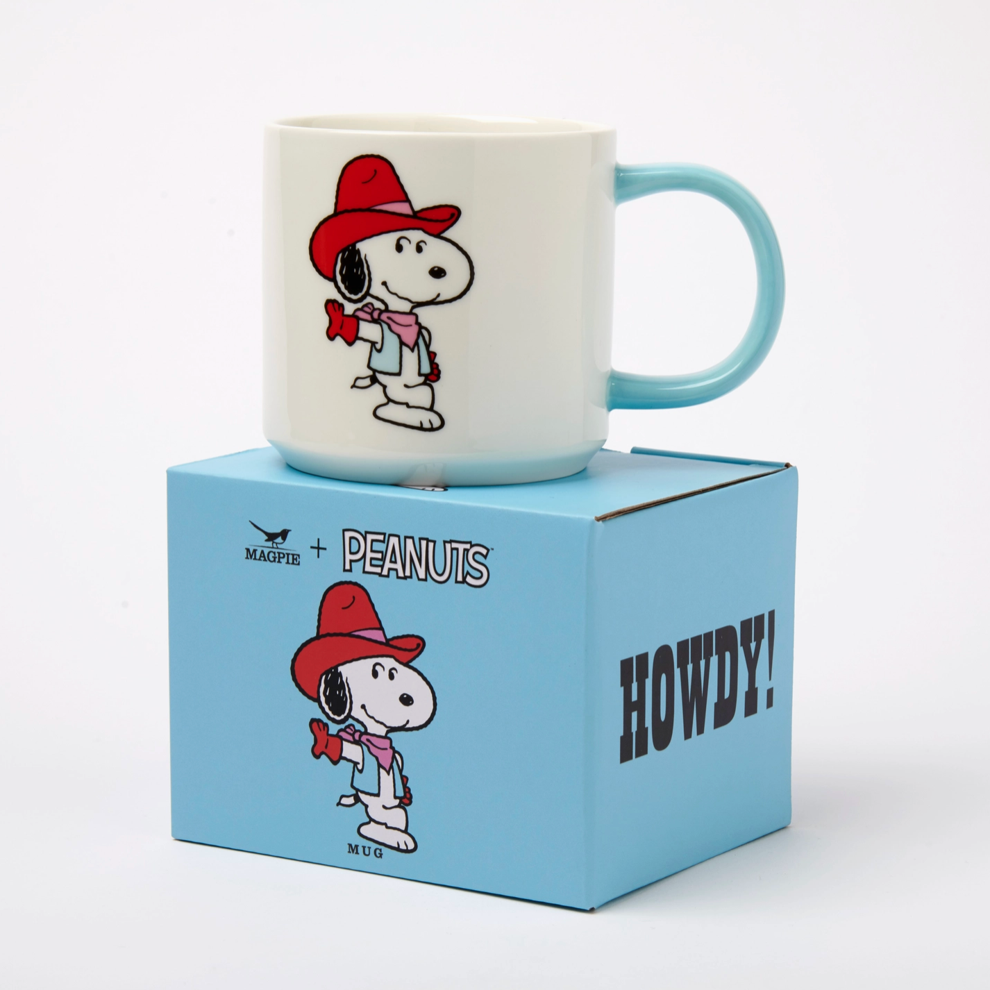 A white ceramic mug with a light blue handle and a printed cartoon character wearing a red hat and pink scarf, displayed on top of a light blue box labeled PEANUTS and HOWDY! against a white studio background.