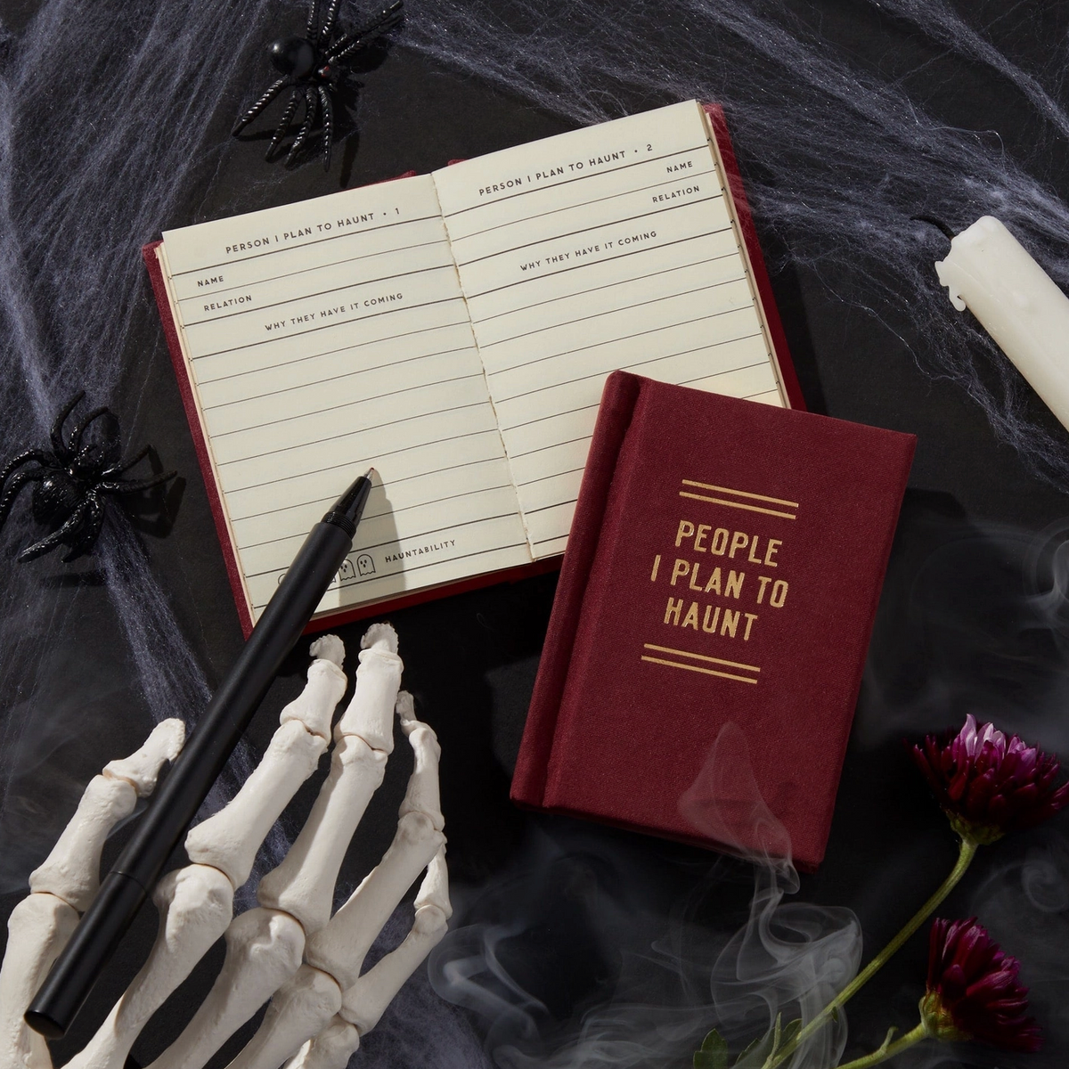 A styled flat-lay featuring a small red hardcover journal with gold foil text reading 'PEOPLE I PLAN TO HAUNT' and an open matching planner notebook. A skeleton hand holds a black pen, with black spiders, cobwebs, a candle, and flowers on a dark smoky background.