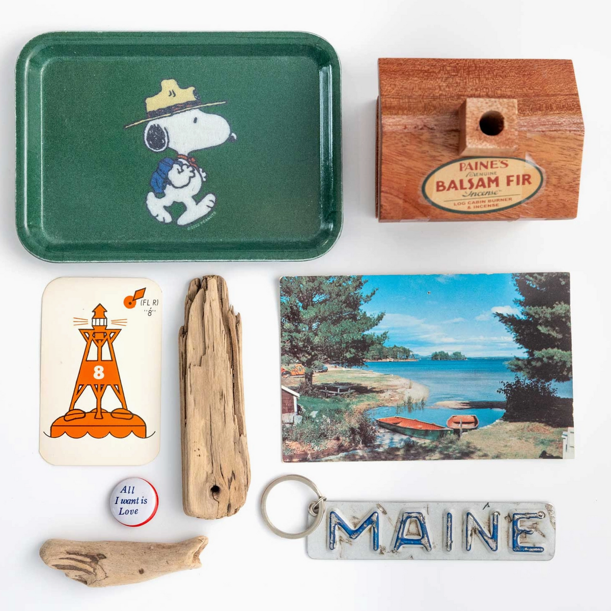 A curated flat-lay collection of vintage and retro items on a white background, including a green Snoopy metal tray, a wooden box of Maine balsam fir, a lighthouse card, driftwood pieces, a hand-shaped keychain, and a Maine state sign.