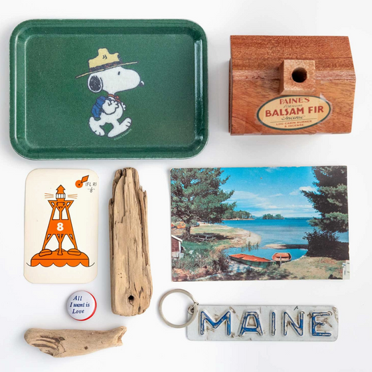 A curated flat-lay collection of vintage and retro items on a white background, including a green Snoopy metal tray, a wooden box of Maine balsam fir, a lighthouse card, driftwood pieces, a hand-shaped keychain, and a Maine state sign.
