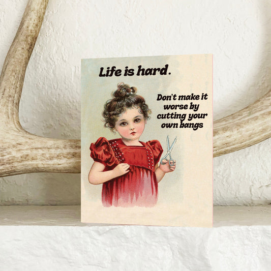 A vintage-style greeting card with a retro illustration of a young girl in a red dress and the text 'Life is hard. Don't make it worse by cutting your own bangs,' displayed on a white shelf in front of antlers.