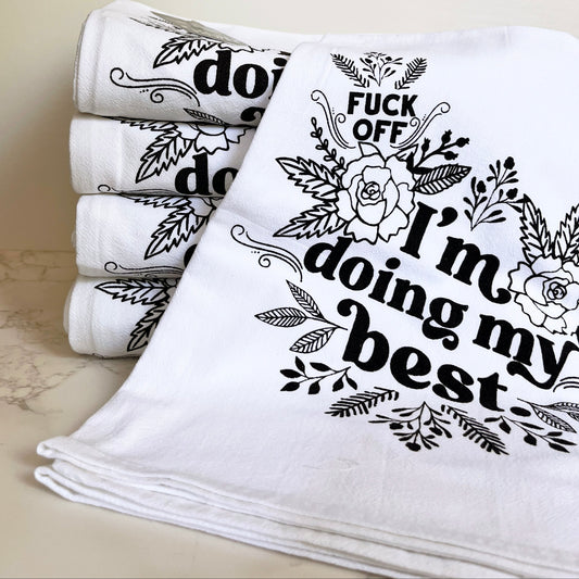 Stack of white towels with black text fuck off im doing my best and floral designs on a light background