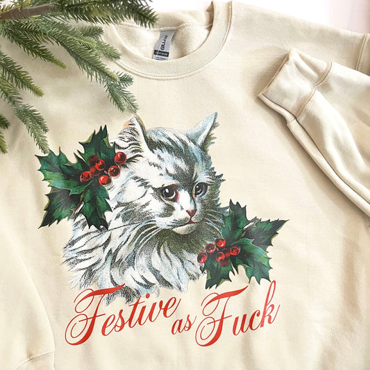A cream-colored sweatshirt with a graphic of a white cat surrounded by mistletoe with the text 'FESTIVE AS FUCK' below it.