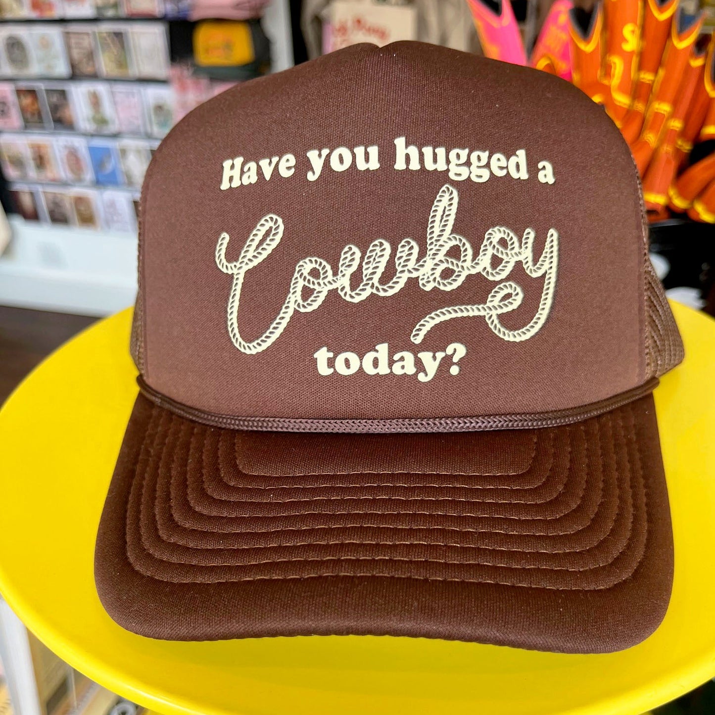 Brown cap with 'Have you hugged a cowboy today?' text on a yellow surface