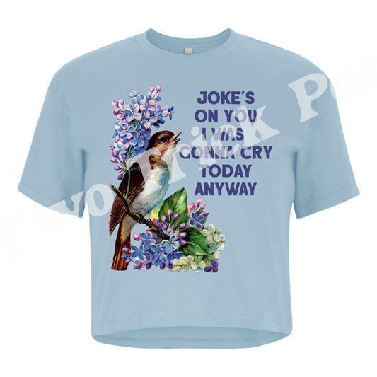 A light blue short-sleeve t-shirt with a graphic print of a kookaburra bird and purple flowers, alongside the text 'JOKE'S ON YOU YOU'RE GONNA CRY TODAY ANYWAY' in purple. The t-shirt is displayed flat against a white background with a faint watermark.