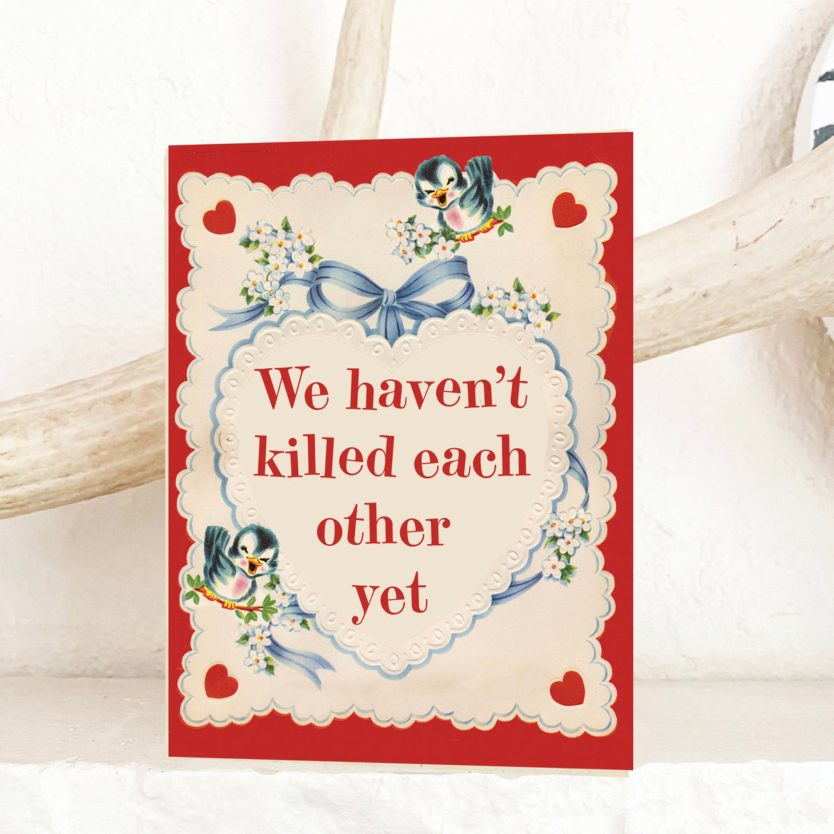 A square greeting card with a red border and cream center panel, featuring penguin illustrations, a blue bow, hearts, and the text 'We haven't killed each other yet.' The card is photographed leaning against a white textured surface with a light wood branch behind it.
