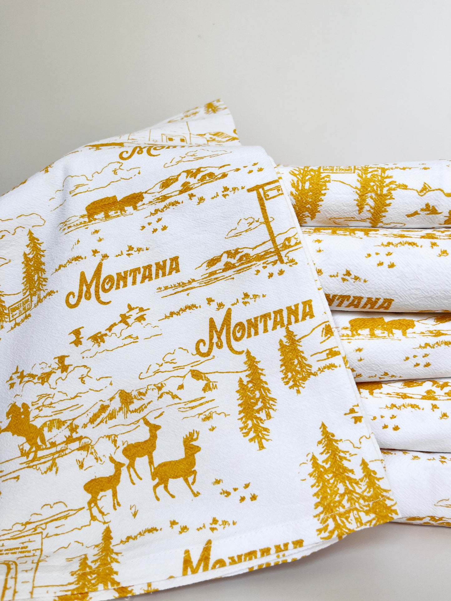 cute cotton tea towel with montana scenes hand drawn illustrations yellow ochre screen print elk bison pine trees ski lift bridger bowl mountains birds roosevelt arch yellowstone glacier national park fun montana souvenir coin laundry cotton kitchen towels 