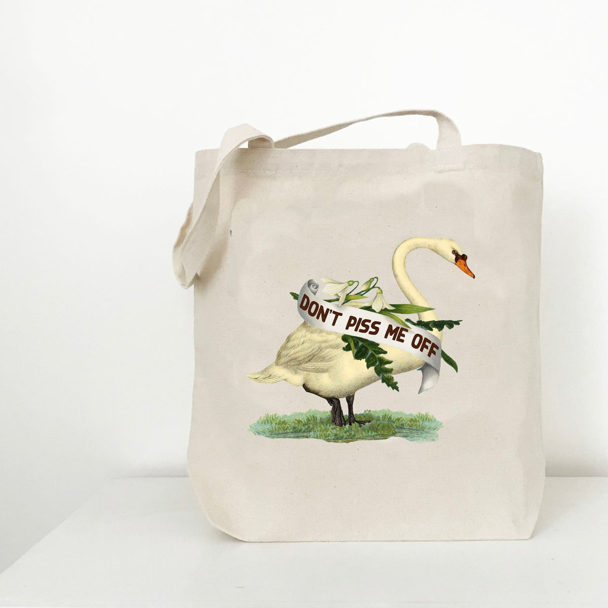 A natural canvas tote bag with a printed graphic of a white swan carrying a banner that reads 'DON'T PISS ME OFF' on a white background.
