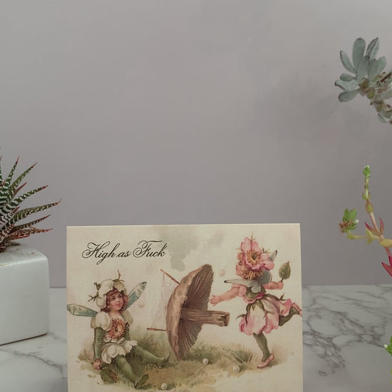 All purpose greeting card featuring a vintage image of two fairies playing badminton, with a mushroom holding the net. One fairy is prancing hitting a ball, while the other fairy is sitting on the ground with a dizzy stare. The greeting reads, "High as Fuck." Vintage pastel colors of pink, green, and cream. Blank inside.  