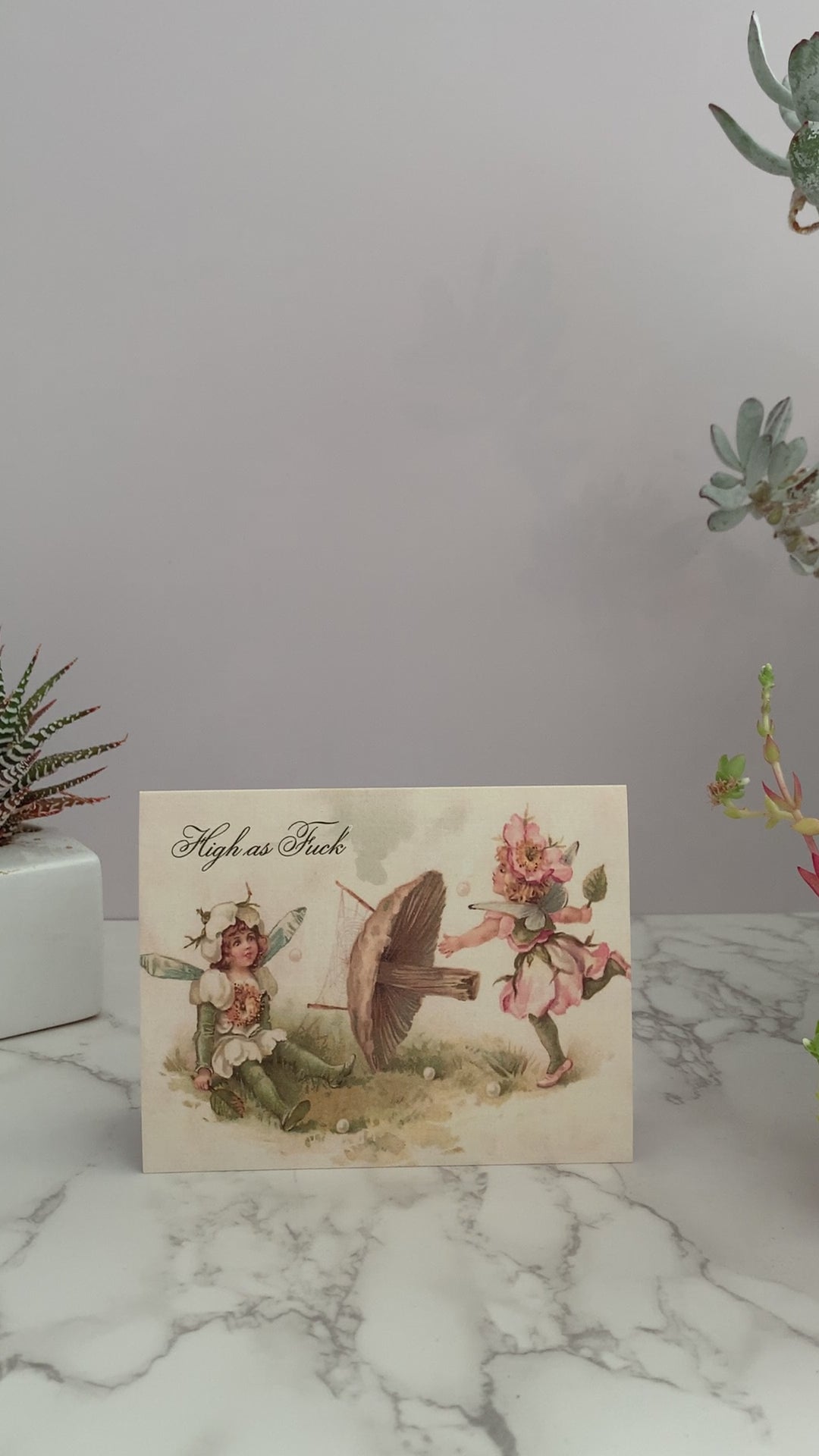 All purpose greeting card featuring a vintage image of two fairies playing badminton, with a mushroom holding the net. One fairy is prancing hitting a ball, while the other fairy is sitting on the ground with a dizzy stare. The greeting reads, "High as Fuck." Vintage pastel colors of pink, green, and cream. Blank inside.  
