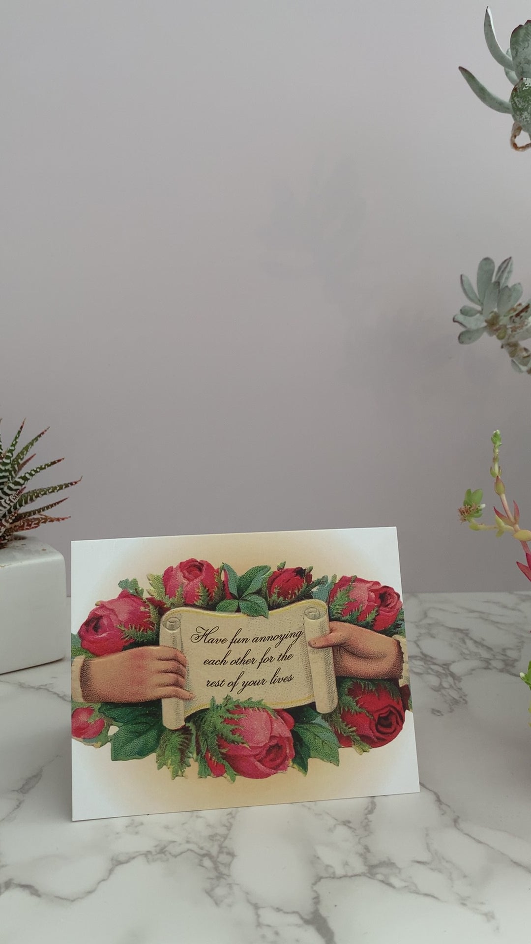 The perfect card for weddings, engagements, and committed partnerships. The front greeting says, "Have fun annoying each other for the rest of your lives." on a paper scroll, being held open by a hand on each side. Red roses surround the image. Vintage inspired. Blank inside.