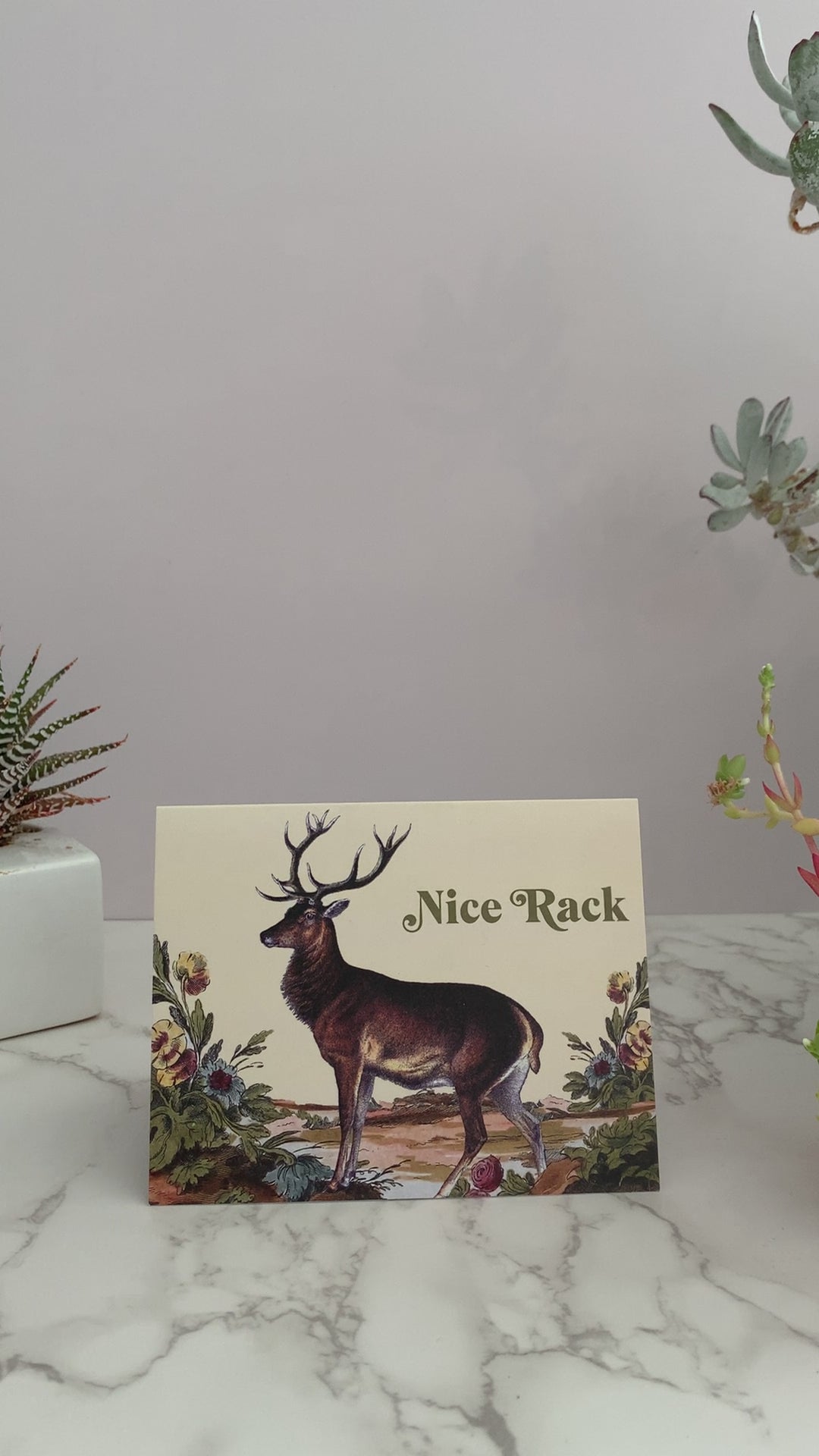 Greeting card featuring a vintage buck deer in an outdoor scene with flowers. Color pallet is vintage yellows, browns, blues, and pinks. Front of card greeting says, "Nick Rack." Great card for any occasion. Send to your crush, your best friend, birthdays, just because. Blank inside. 