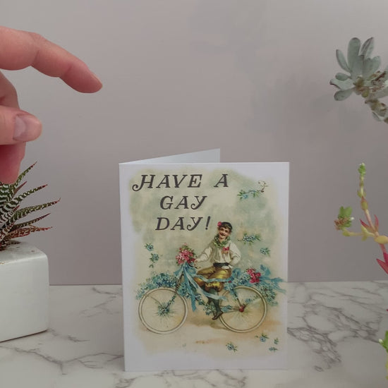 All purpose greeting card that says "Have a gay day!" on the front. Vintage image featuring a happy man with a mustache on a bicycle, with flowers and ribbons. Color pallet is yellows and blues with a little pink. Blank inside.