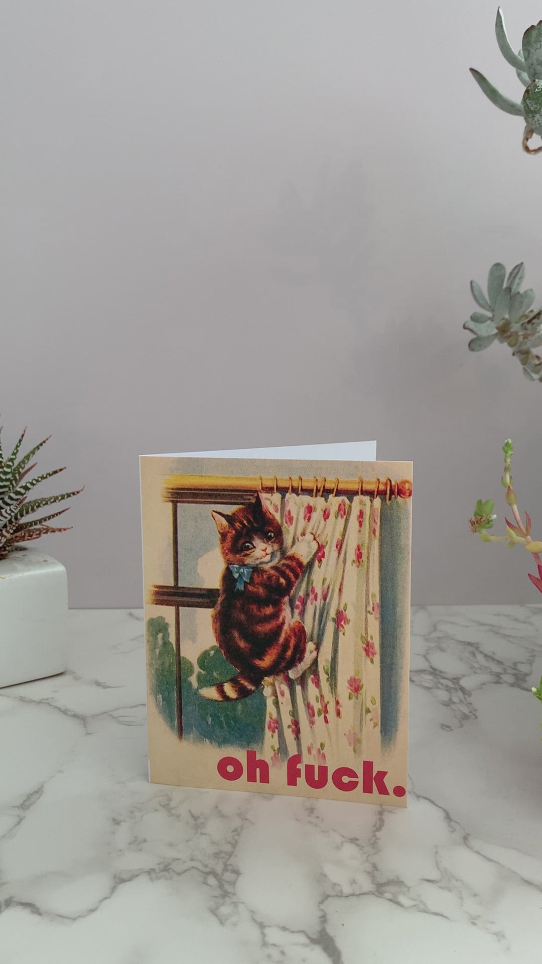 Greeting card with a vintage image of a brown and orange stripped kitty that has climbed up floral drapes covering a window. Greeting says, "Oh fuck." Color pallet is vintage yellows, cream, blues, and pinks. Blank inside. 