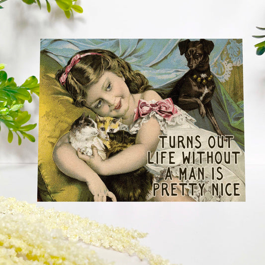 A square greeting card displayed on a white surface, featuring a vintage-style illustration of a young girl hugging a cat, with a dog in the background and the text 'TURNS OUT LIFE WITHOUT A MAN IS PRETTY NICE'. The scene is styled with green leaves and pale flowers around it.